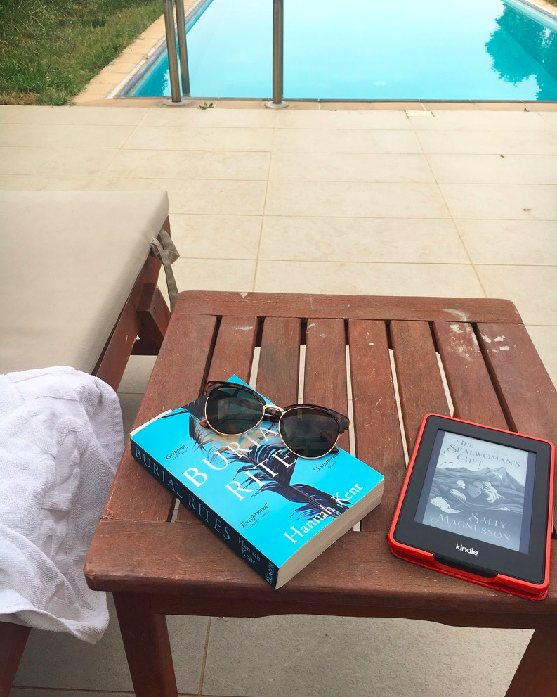 Burial Rites by Hannah Kent and The Sealwoman's Gift by Sally Magnusson laying upon a table by a sun lounger, next to a pool in Greece.